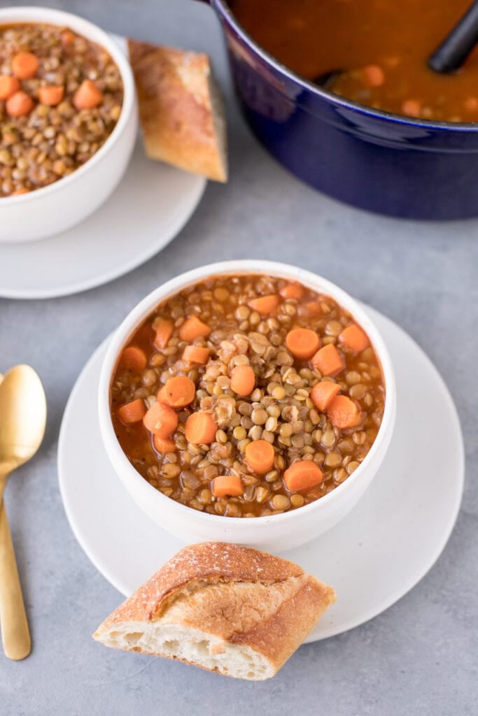 Two white bowl of easy lentil soup with a gold spoon and bread on the side with a pot of lentil soup on the side