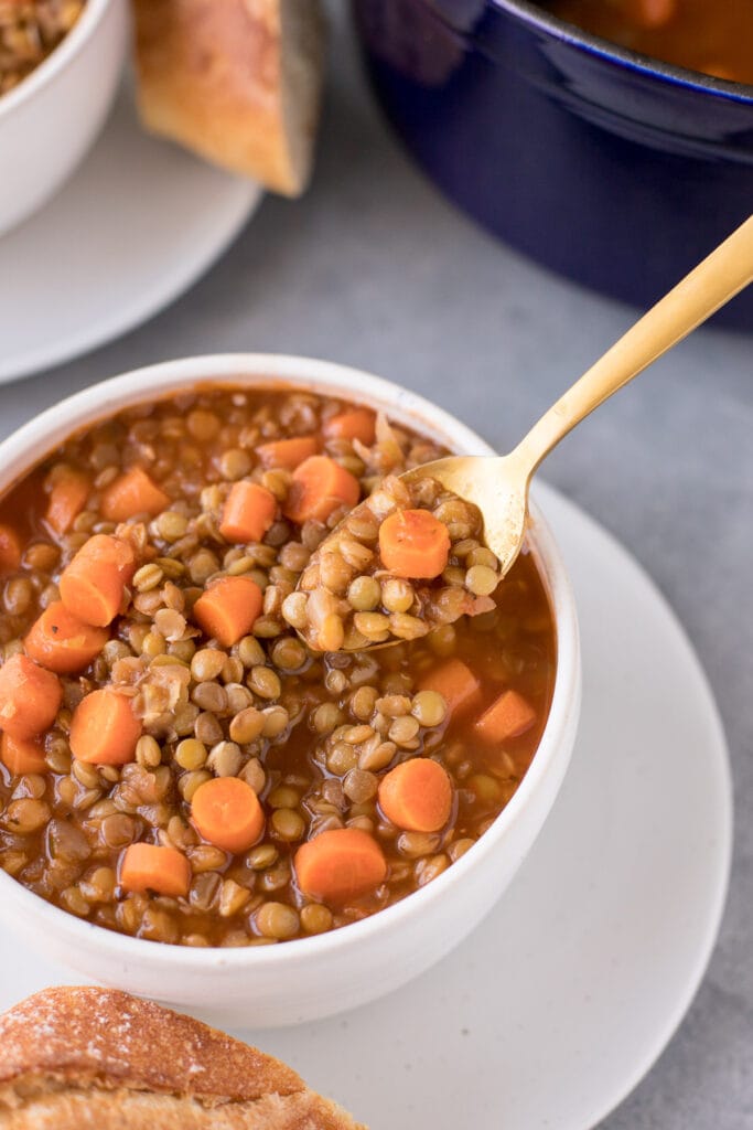 A bowl of easy lentil soup on a white plate with bread off to the side and a bite being taken out of it with a gold spoon