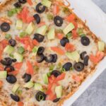 Taco Dip in a white dish on a grey background with toppings on it (olives, tomatoes, avocados)
