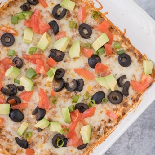 Taco Dip in a white dish on a grey background with toppings on it (olives, tomatoes, avocados)
