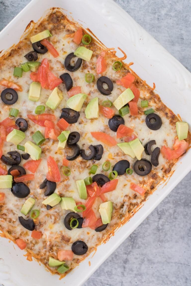 Taco Dip in a white dish on a grey background with toppings on it (olives, tomatoes, avocados)