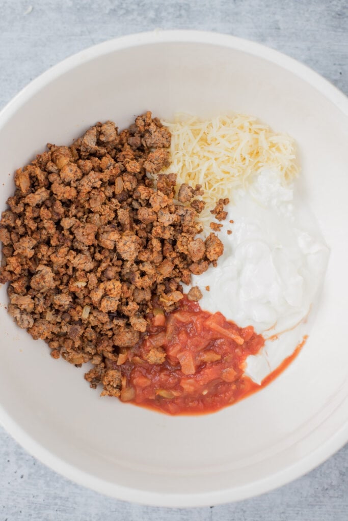 Cooked ground beef, salsa, cottage cheese, greek yogurt and cheddar cheese in a bowl before getting mixed together for taco dip
