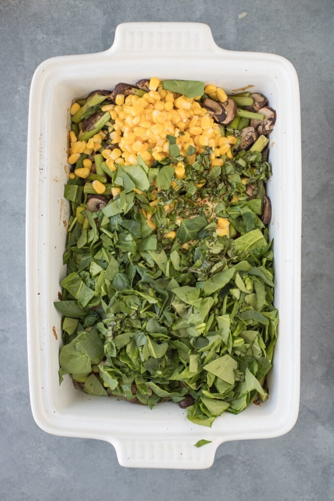 corn and spinach being added on top of baked feta and vegetables for feta pasta