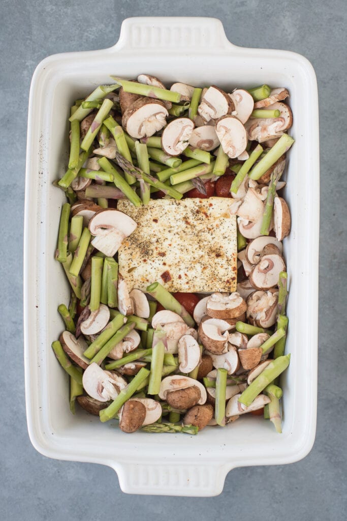 A white baking dish with tomatoes and feta. Asparagus and mushrooms have been placed around the feta on top of the tomatoes