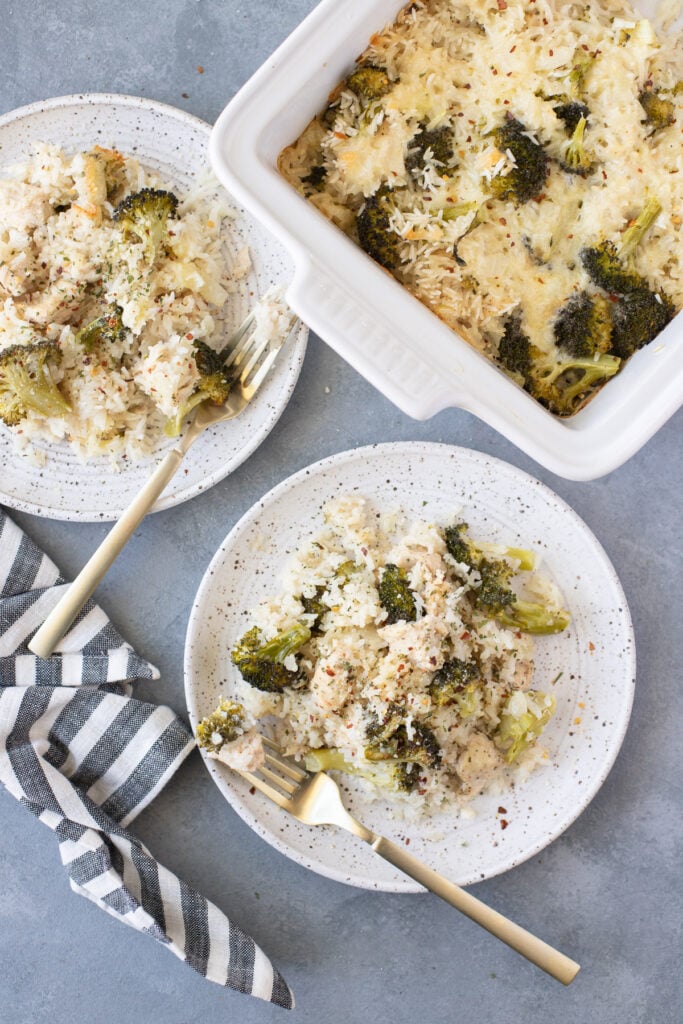 An overhead shot of two white plates with cheesy chicken and rice casserole with gold forks and a casserole dish of the chicken and rice casserole on a grey background