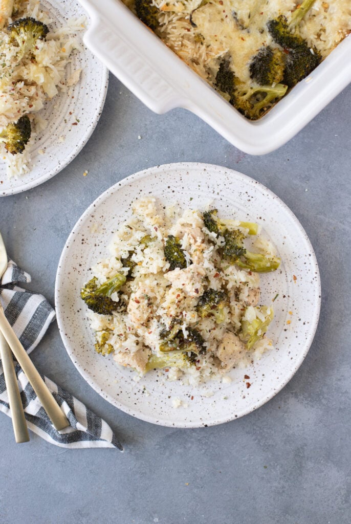 An overhead shot of a white plate with cheesy chicken and rice casserole with gold forks on the side and a white casserole dish of the chicken and rice casserole on a grey background