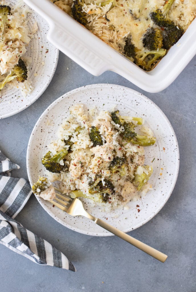 An overhead shot of a white plate with cheesy chicken and rice casserole with gold forks and a white casserole dish of the chicken and rice casserole on a grey background