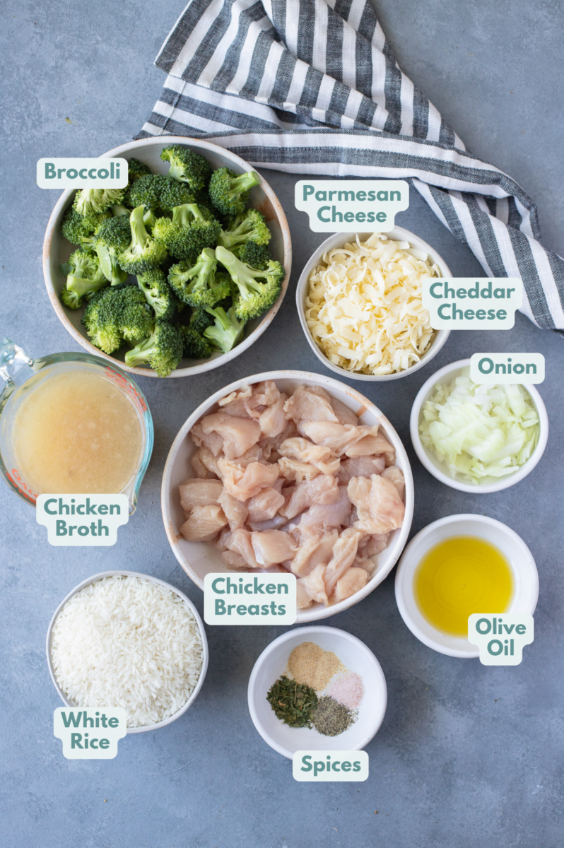 Ingredients for chicken and rice casserole in small white bowls on a grey background: chicken breasts, brown rice, olive oil, chicken broth, broccoli florets, onion, pepper, garlic powder, parsley, salt, parmesan cheese, cheddar cheese