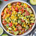 Corn Tomato Avocado Salad in a white bowl on a grey background with tomatoes, avocado and limes off to the side