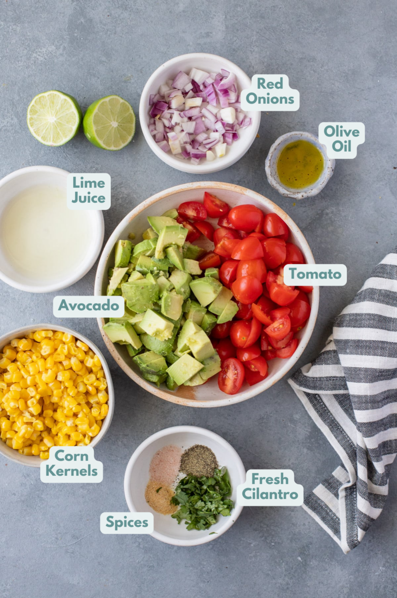 Ingredients of Corn Tomato Avocado Salad in small white bowls on a grey background: avocado, grape tomatoes, fresh corn, red onion, extra virgin olive oil, salt and pepper, garlic powder, fresh lime juice, fresh cilantro