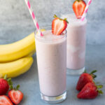 Two Strawberry Banana Smoothies in glass jars on a grey background with strawberries and bananas off to the side