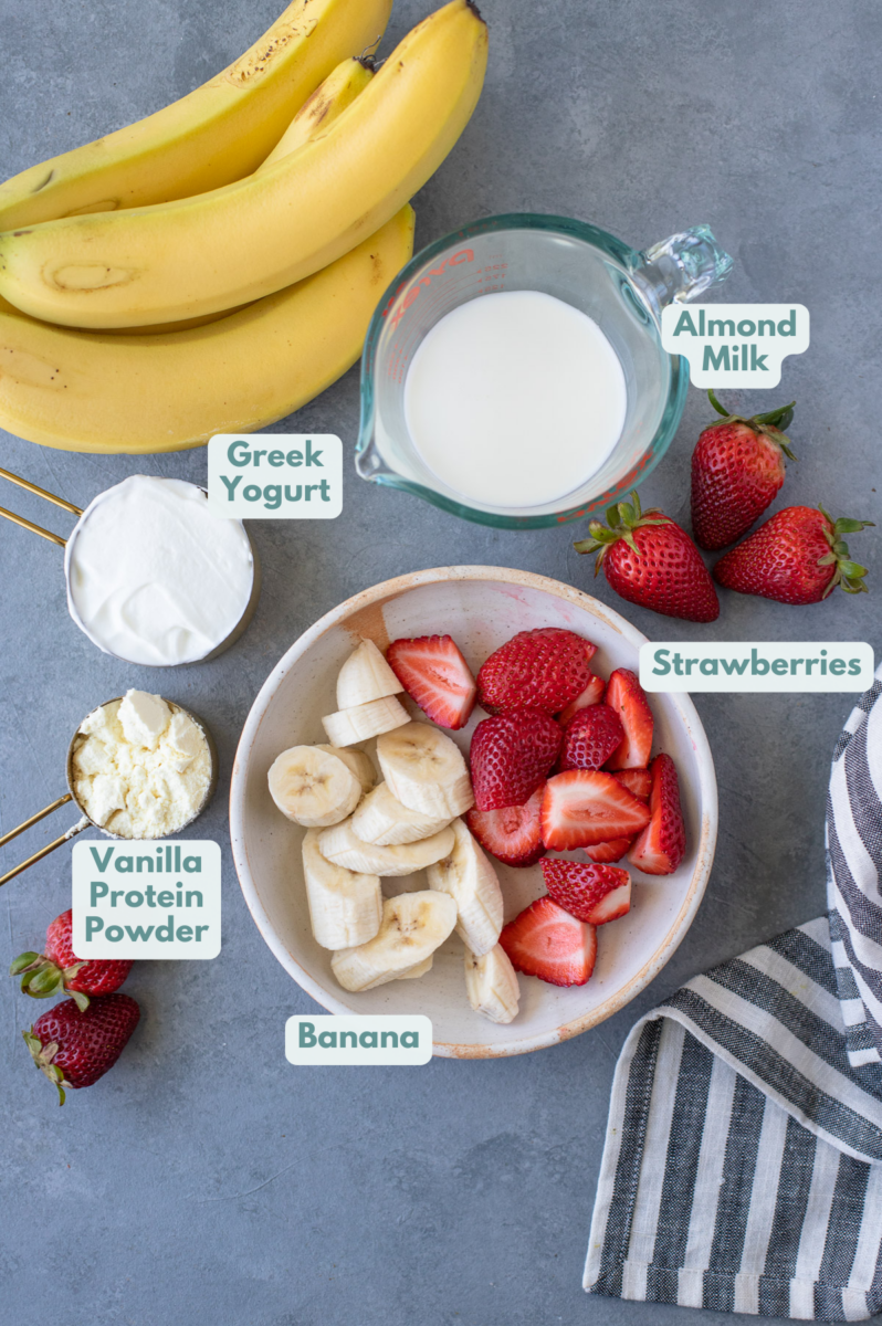 Ingredients for a Strawberry Banana Smoothie on a grey background: strawberries, bananas, protein powder, greek yogurt and almond milk