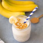 The Best Banana Smoothie Recipe on a grey background with bananas and peanut butter in the background