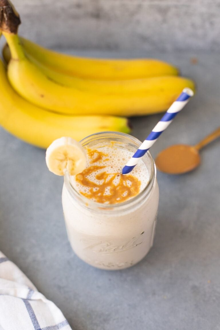 The Best Banana Smoothie Recipe on a grey background with bananas and peanut butter in the background