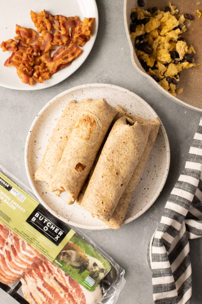 Rolled up breakfast burritos on a plate with bacon and scrambled eggs off to the side