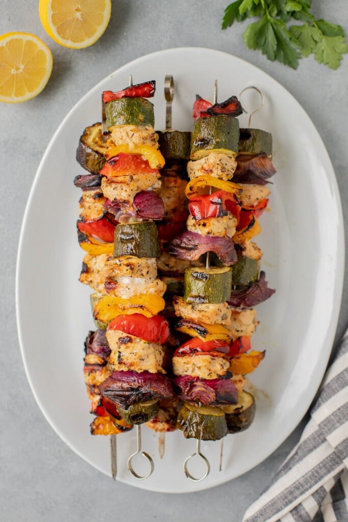 Greek Chicken Kabobs Recipe on a white platter after coming off of the grill with lemons and fresh herbs on the side.
