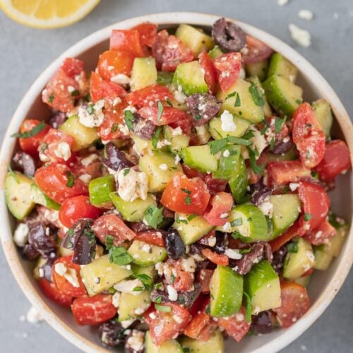 A bowl of traditional greek salad on a grey background with ingredients all around it