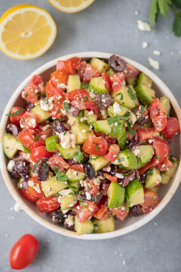 A bowl of traditional greek salad on a grey background with ingredients all around it