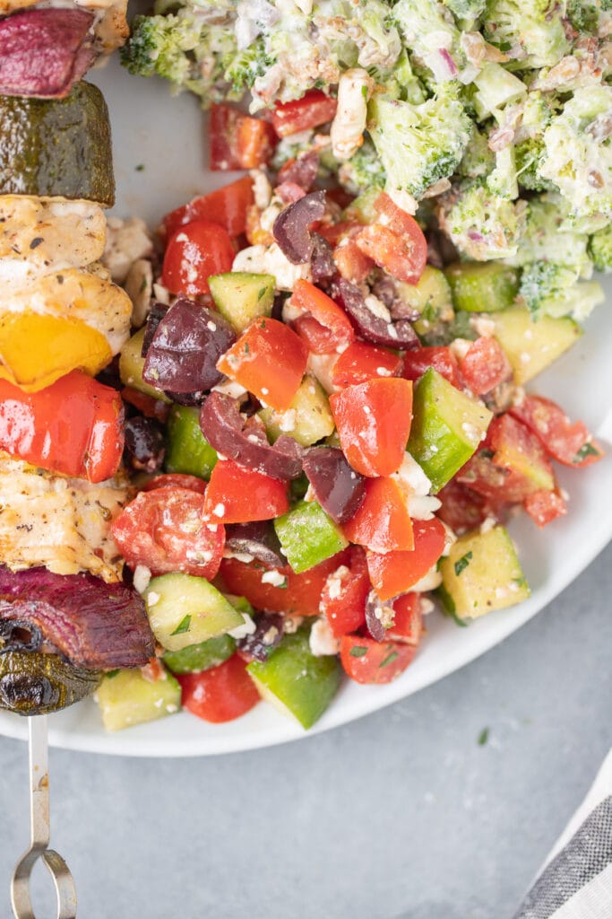 A close up of greek salad on a white plate with kebabs and broccoli salad off to the side