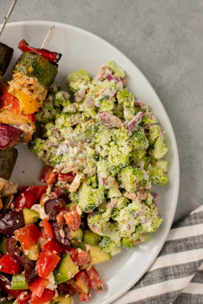 The best broccoli salad recipe on a plate with greek salad and chicken kababs