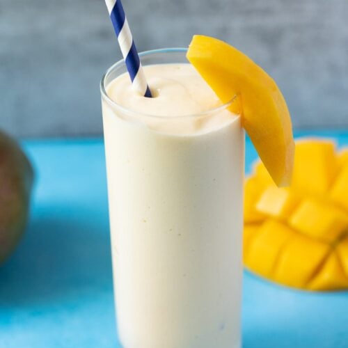 A mango smoothie on a blue background with mangos off to the side and a blue paper straw