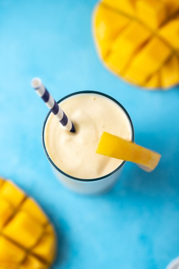 An overhead shot of a mango smoothie recipe on a bright blue background with sliced mangoes off to the sides.