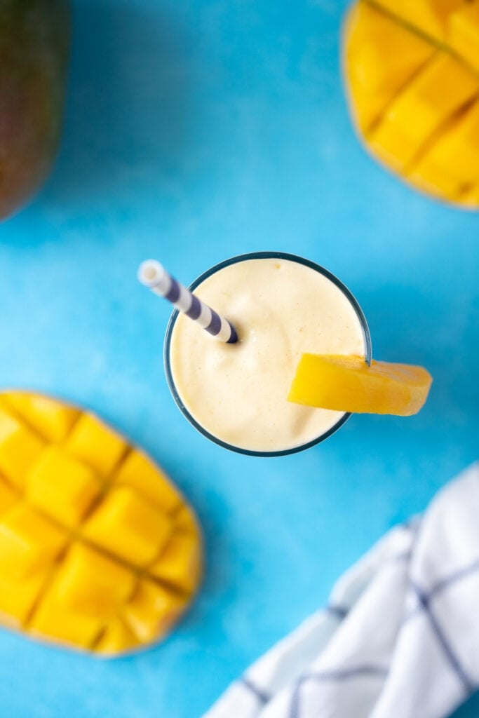 An overhead shot of a mango smoothie on a blue background with cut and whole mangos off to the side
