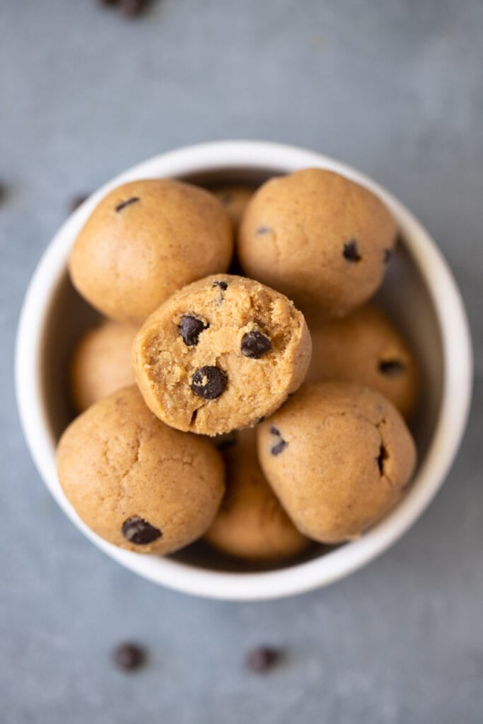 A close up of no bake protein balls with a bite taken out of them in a small white bowl on a grey background with chocolate chips off to the side