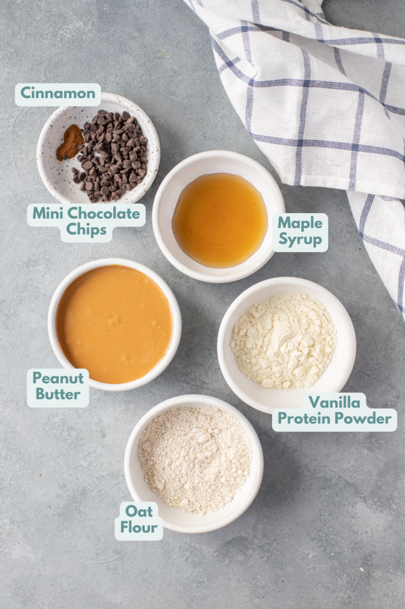Ingredients for no bake protein balls in small white bowls on a grey background with a checked linen off to the side