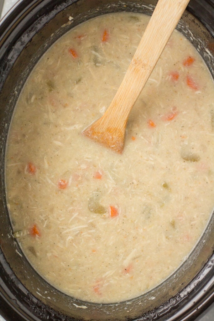 a close up of chicken pot pie soup in a crockpot with a wooden spoon stirring it