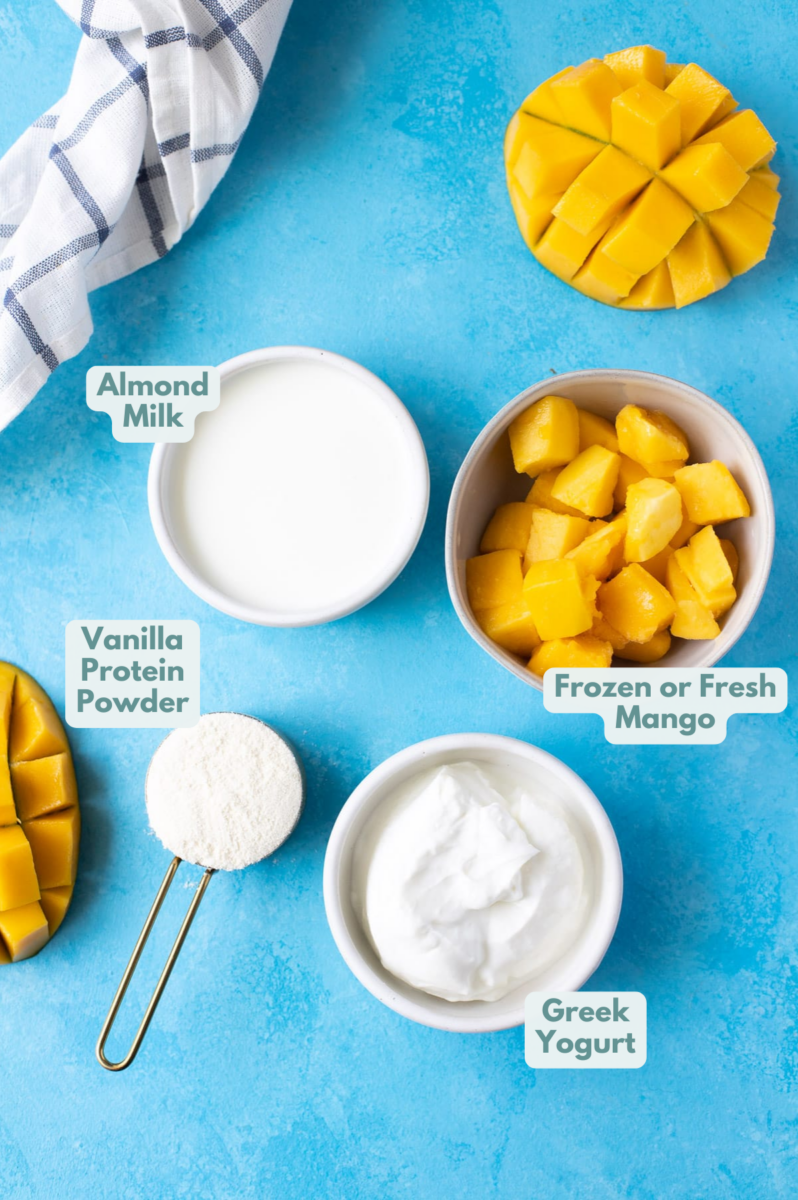 Ingredients for Mango Smoothie in small white bowls on a blue background: greek yogurt, almond milk, mango and protein powder