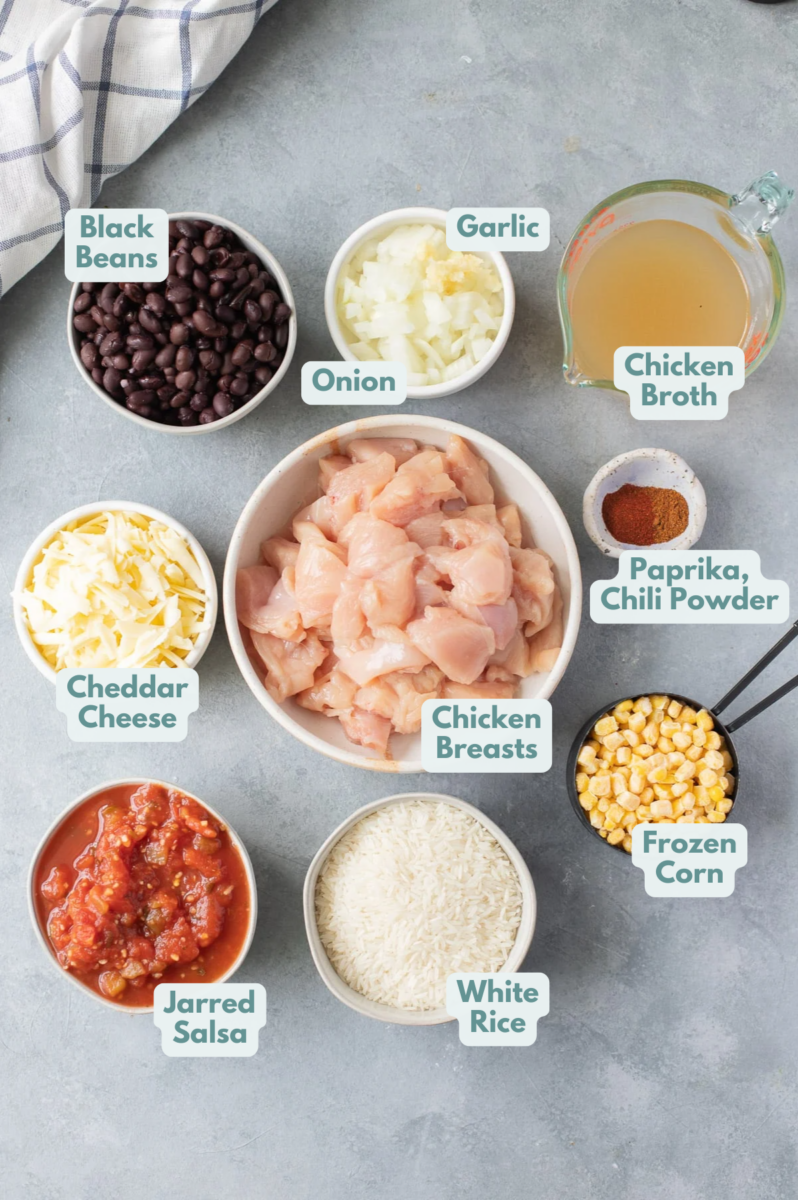 Ingredients for Southwest Chicken Casserole in small white bowls: white rice, minced garlic, chopped onion, chicken broth, chicken breasts, black beans, frozen corn, jarred salsa, chili powder, paprika, and cheddar cheese