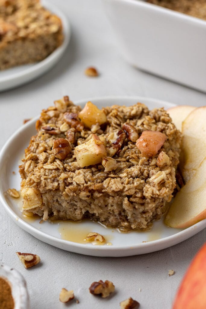 A close up of a slice of healthy baked oatmeal on a white plate with a baking dish off to the side and nuts scattered around the dish