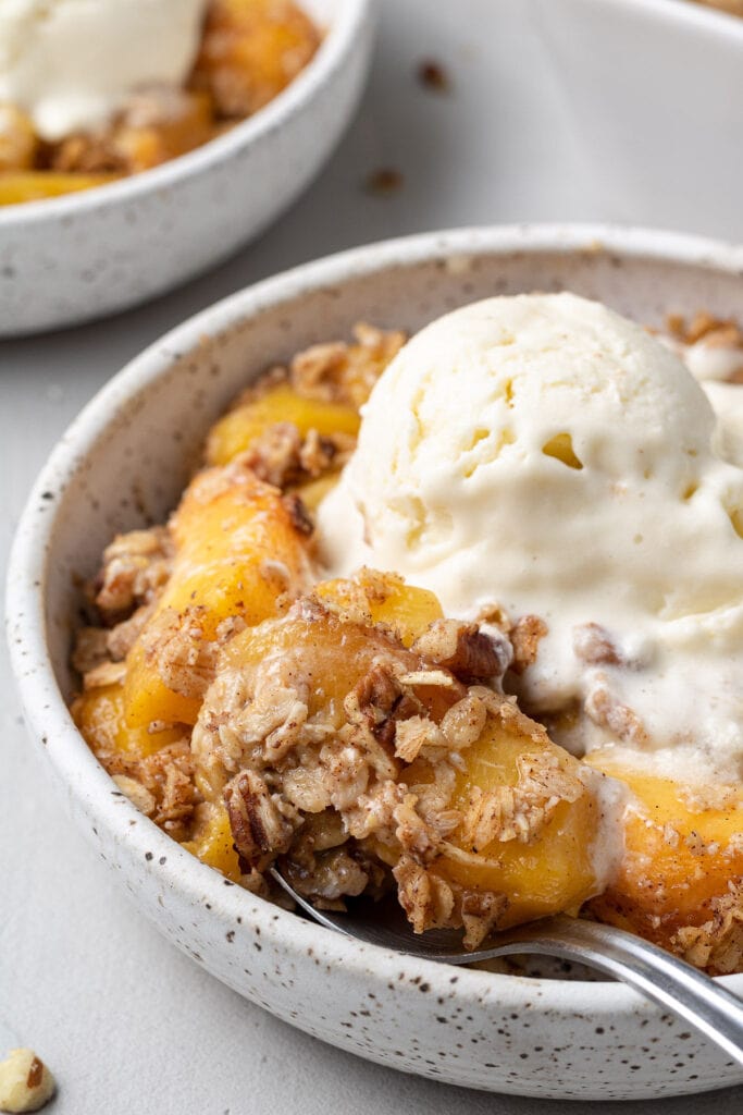 A bowl of healthy peach crisp with a spoon taking a bite out of it.