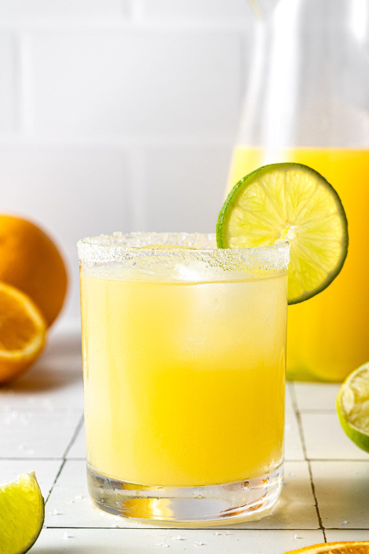 A cold skinny margarita in a salt-rimmed, short cocktail glass with oranges and limes and a pitcher of skinny margaritas off to the side
