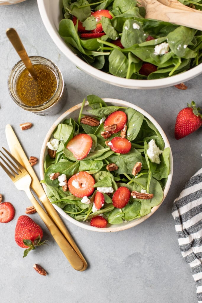 a bowl of strawberry spinach salad on a grey background with a bowl off to the side