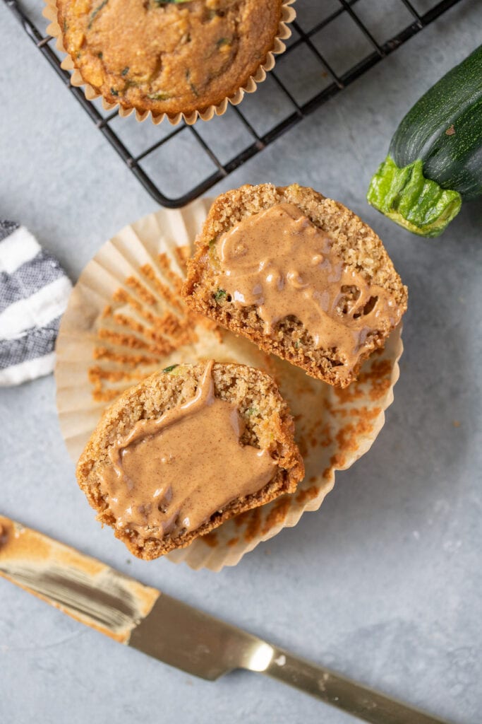 A healthy zucchini muffin sliced in half with peanut butter spread on it with a knife, zucchini and another muffin on a cooling rack surrounding it all on a grey background