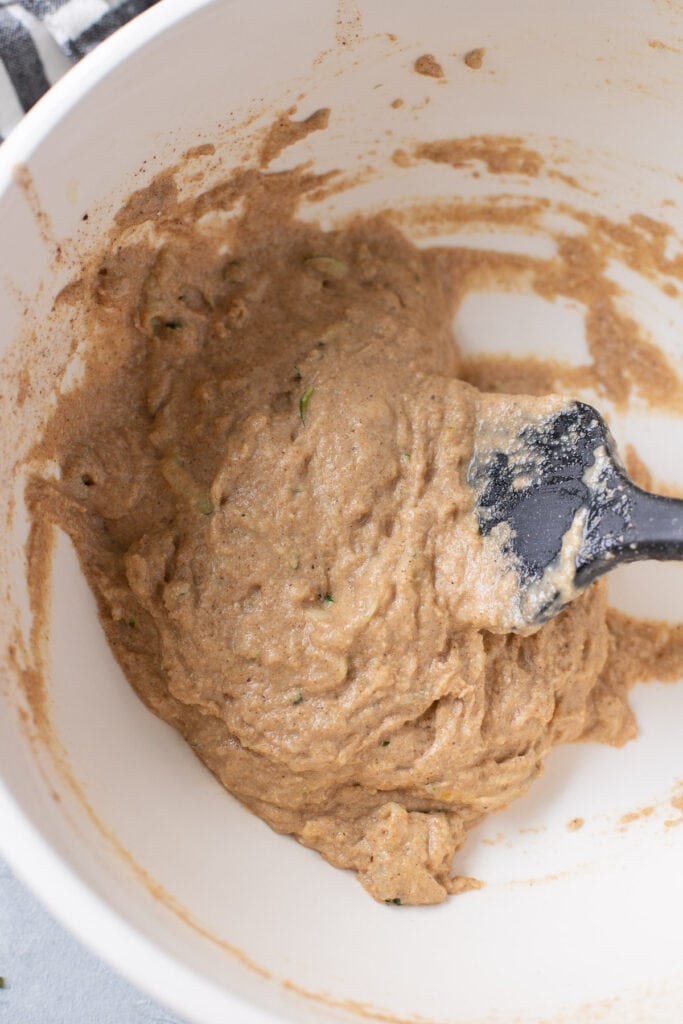 Batter for healthy zucchini muffins being stirred together with a spatula in a white bowl
