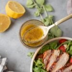 A close up of a spoon dipping out some of the best Greek Salad Dressing with lemon, herbs and bbq chicken salad on the side on a grey background