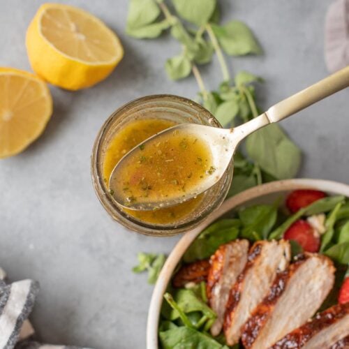 A close up of a spoon dipping out some of the best Greek Salad Dressing with lemon, herbs and bbq chicken salad on the side on a grey background