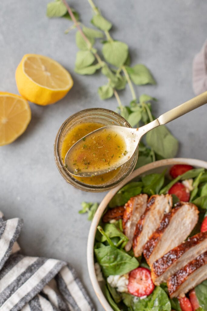 A close up of a spoon dipping out some of the best Greek Salad Dressing with lemon, herbs and bbq chicken salad on the side on a grey background