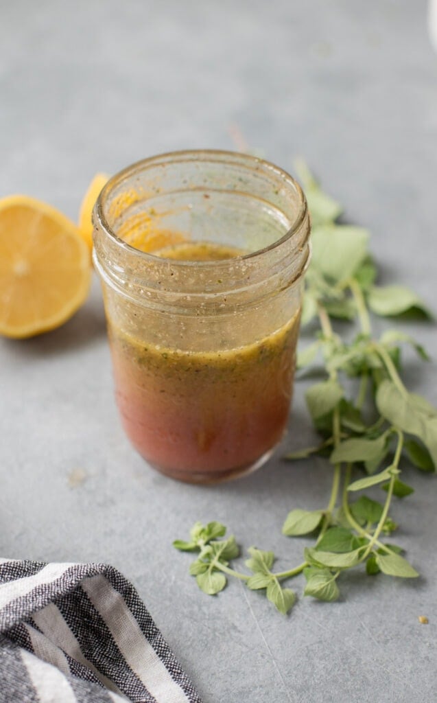 A close up of Healthy Greek Salad Dressing with lemon and herbs on the side on a grey background