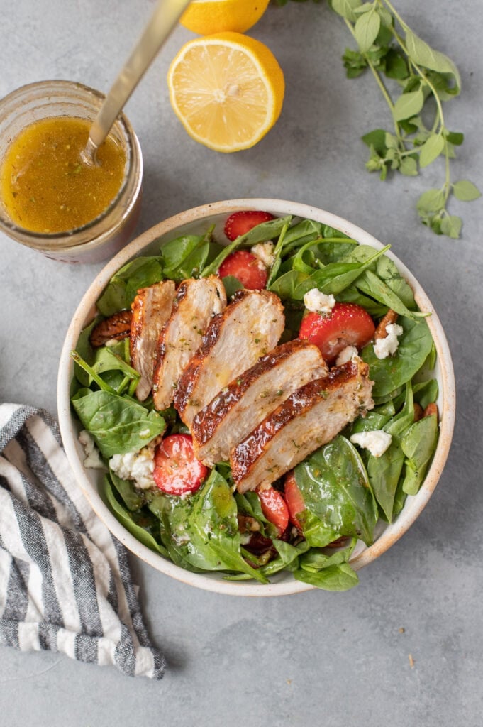 A close up of grilled BBQ Chicken sliced on a strawberry spinach salad with traditional Greek salad dressing on the salad and a jar on the side with lemon and herbs on the side and feta cheese and grilled chicken on the salad