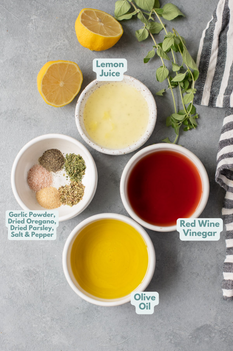 ingredients for the Homemade Greek Dressing - red wine vinegar, lemon juice, olive oil, garlic powder, dried oregano, dried parsley, salt, pepper in white bowls on a grey background