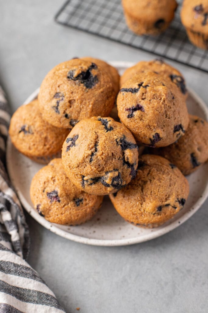 A close up of Blueberry Muffins on a white plate on a grey background with a striped linen off to the side