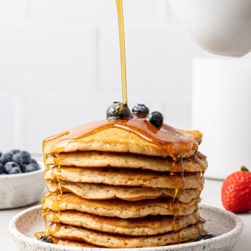a stack of fluffy healthy pancakes with blueberries on top and syrup being poured over it