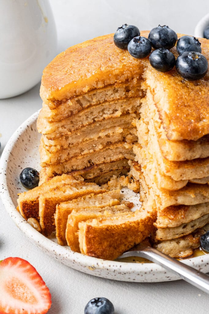 a close up of a stack of fluffy healthy pancakes with blueberries on top and with a bite taken out of them
