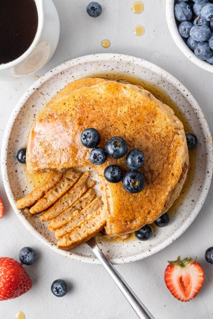 an overhead shot of a stack of fluffy healthy pancakes with blueberries on top and with a bite taken out of them