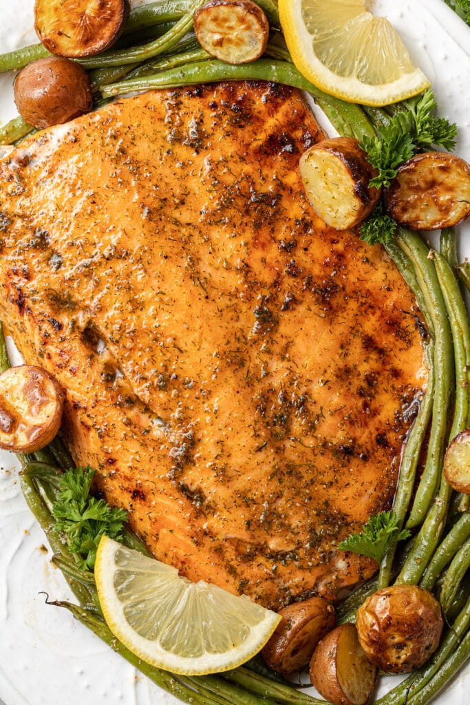 A close up of lemon salmon on a white dish with green beans, potatoes and lemon slices on the side