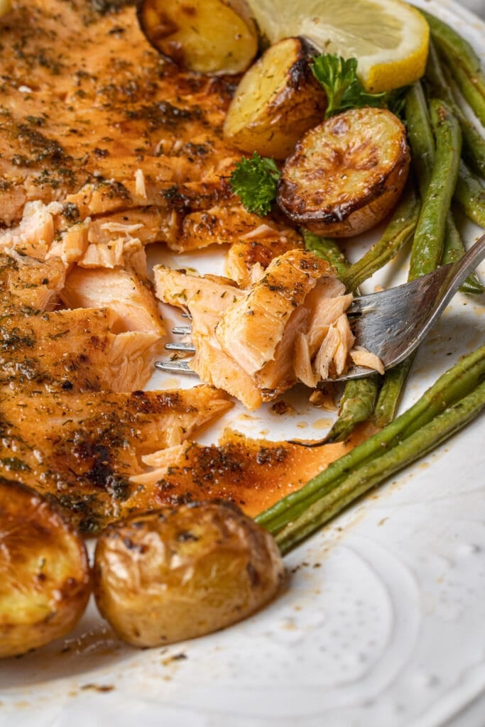 A close up of flaky lemon salmon on a white dish fresh out of the oven with a lemon slice on the side